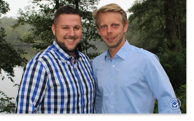 Two men standing side by side outdoors by a lake, smiling at the camera with trees in the background.